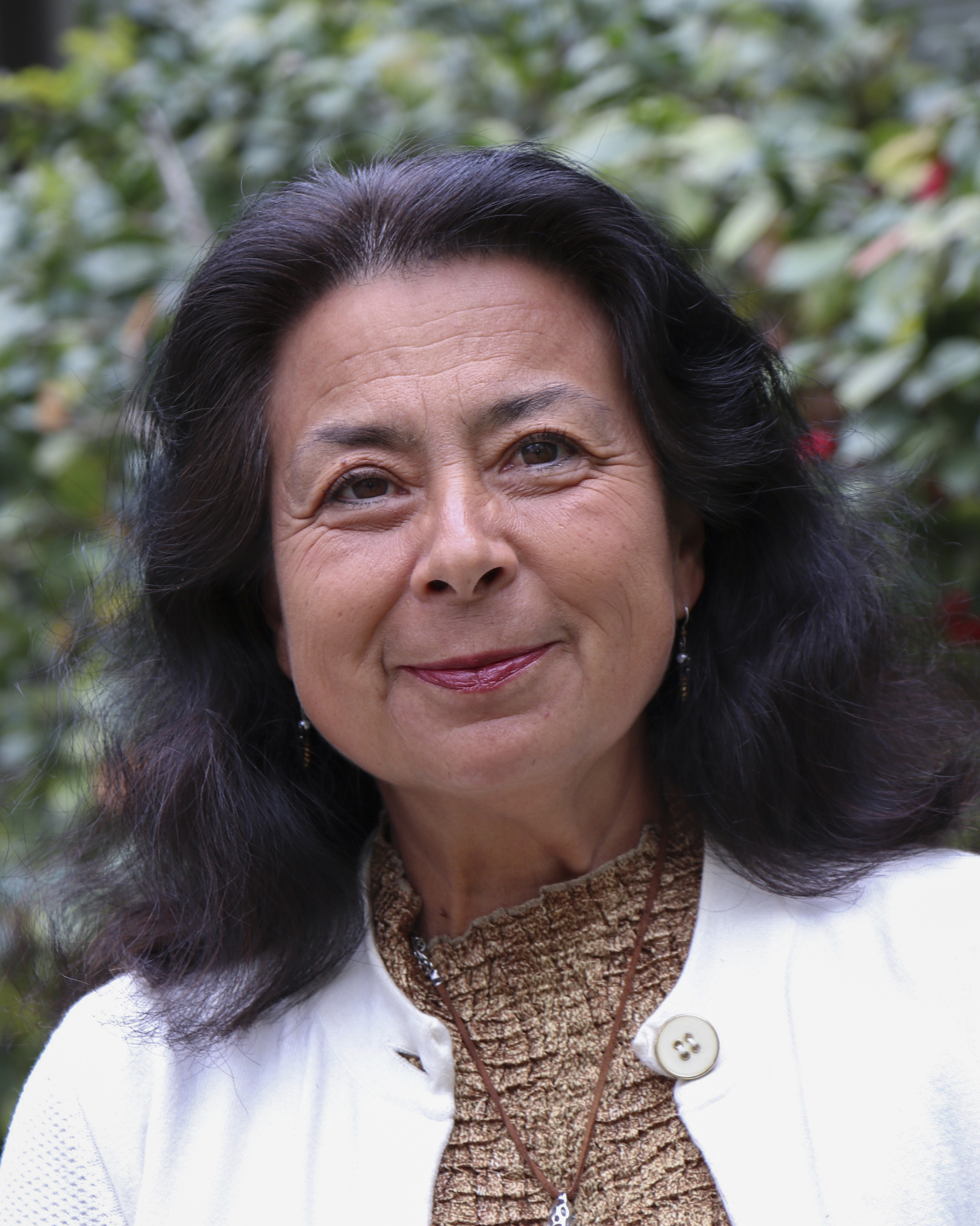 Headshot of Dr. Rose Borunda. Dr. Borunda stands in front of a dark green bush with red flowers. Dr. Borunda smiles wearing a white cardigan and bown top